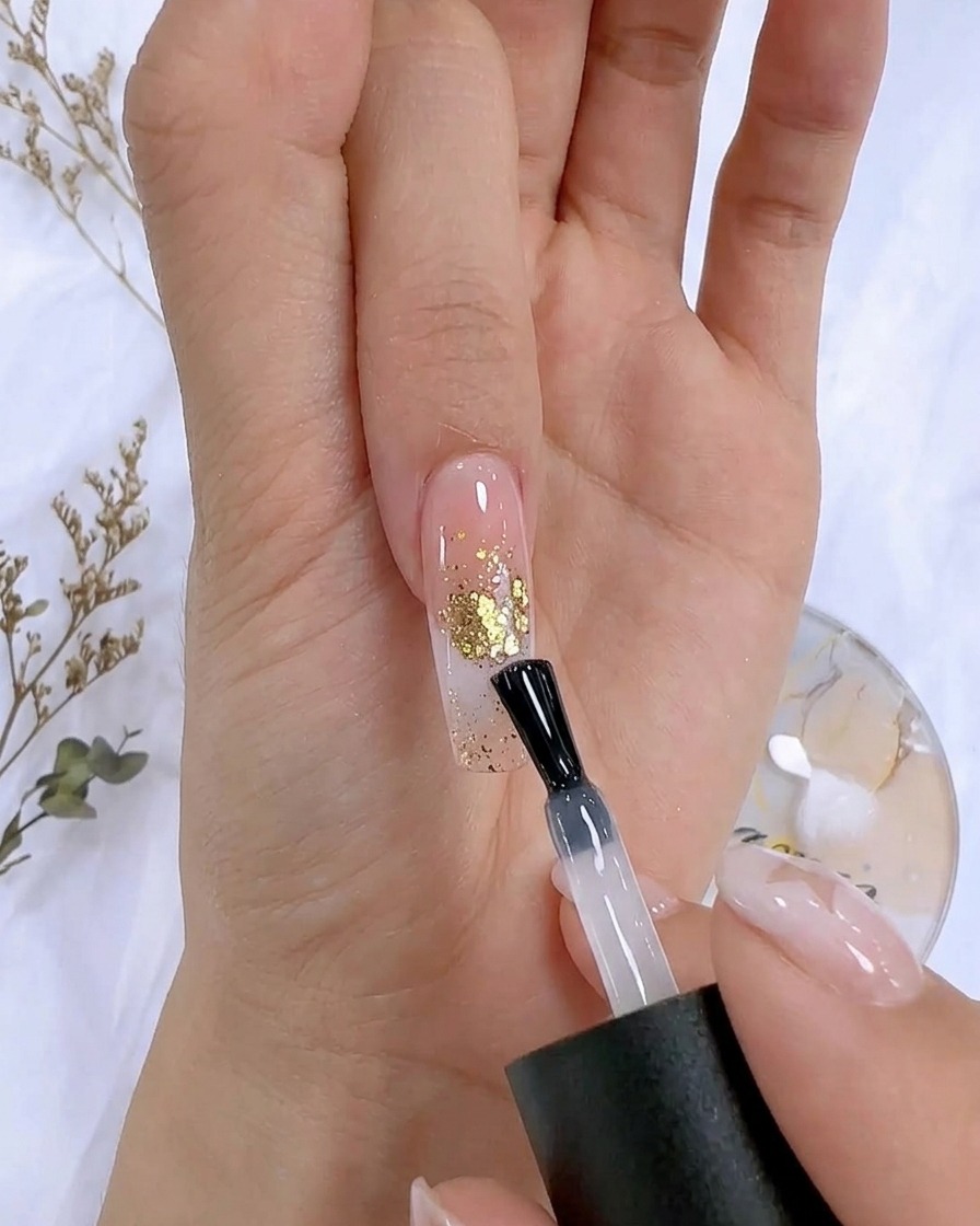 Un pinceau à vernis appliquant un top coat transparent sur un ongle décoré daccents en feuille dor.