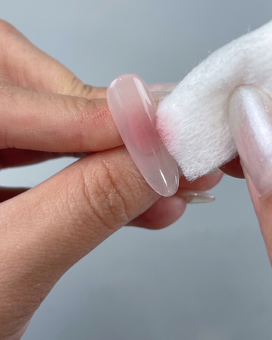 Une main utilisant un bloc-éponge blanc pour tamponner du vernis gel rose blush sur le centre dun ongle en amande transparent.