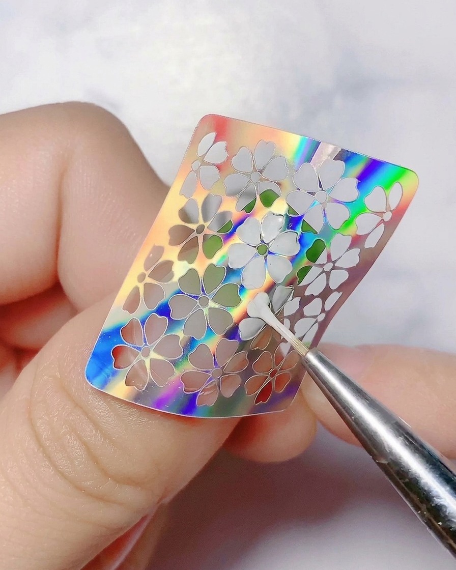 Un outil fin retirant délicatement un sticker de fleur holographique de longle.