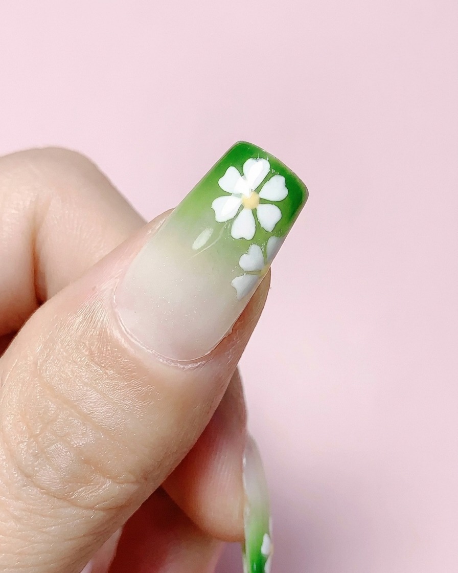 Un ongle carré terminé avec un dégradé vert et des fleurs blanches, recouvert dun top coat transparent.