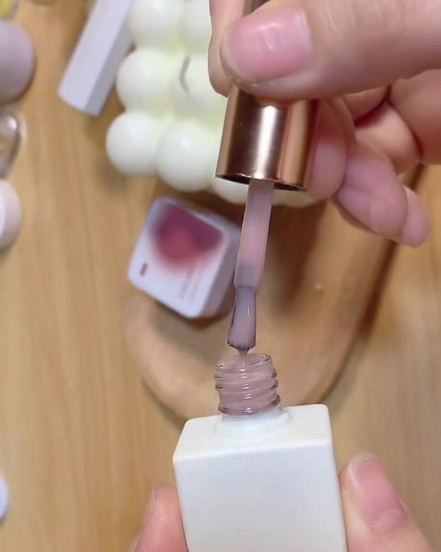 Un pinceau appliquant une base rose translucide depuis un flacon de vernis blanc.