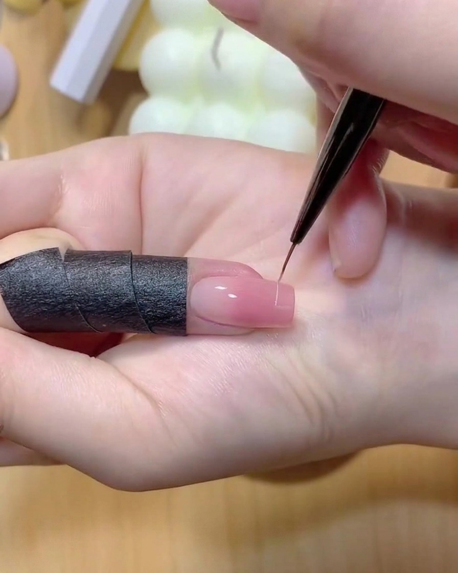Un pinceau fin dessinant une fine ligne blanche sur le bout dun ongle dégradé rose.