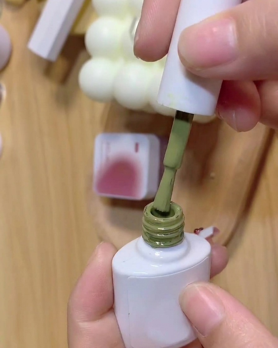 Un pinceau trempant dans un flacon de vernis gel vert avec un pot de gel rose en arrière-plan.