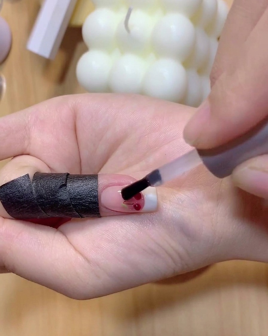 Un pinceau appliquant du vernis gel rouge translucide sur les boules de cerise 3D transparentes.