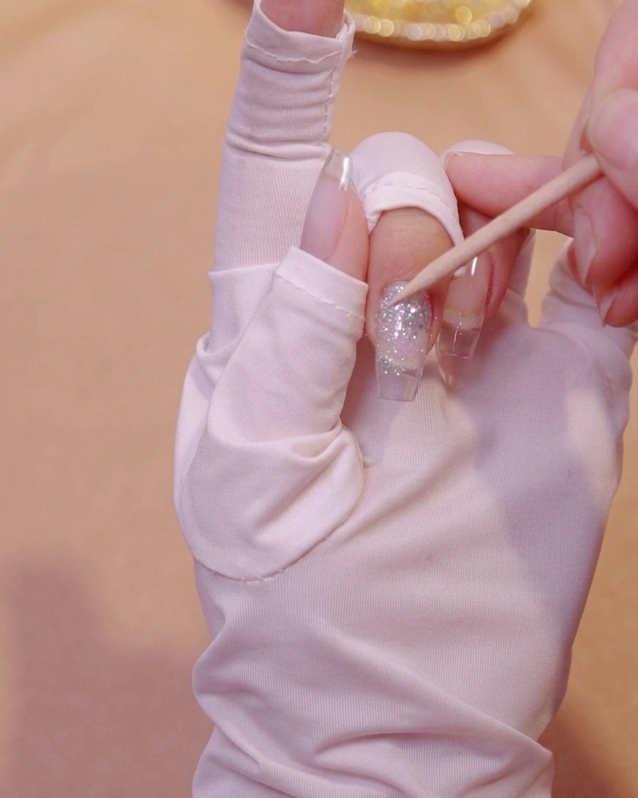 Un bâtonnet de buis appuyant doucement sur des sequins papillons incrustés dans du gel pailleté sur un ongle.