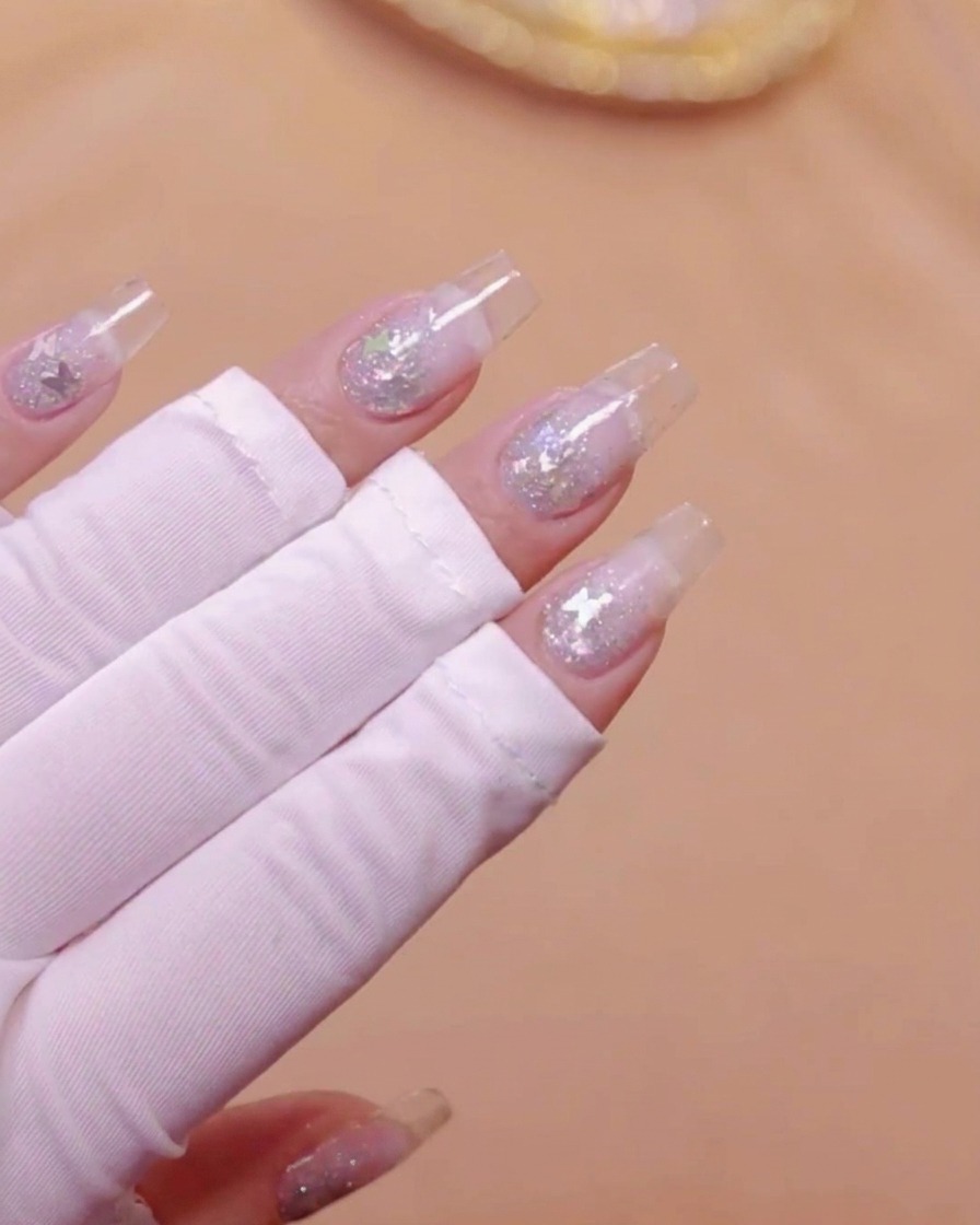 Une main avec des extensions dongles transparentes et des paillettes papillons incrustées, positionnée sous une lampe UV.