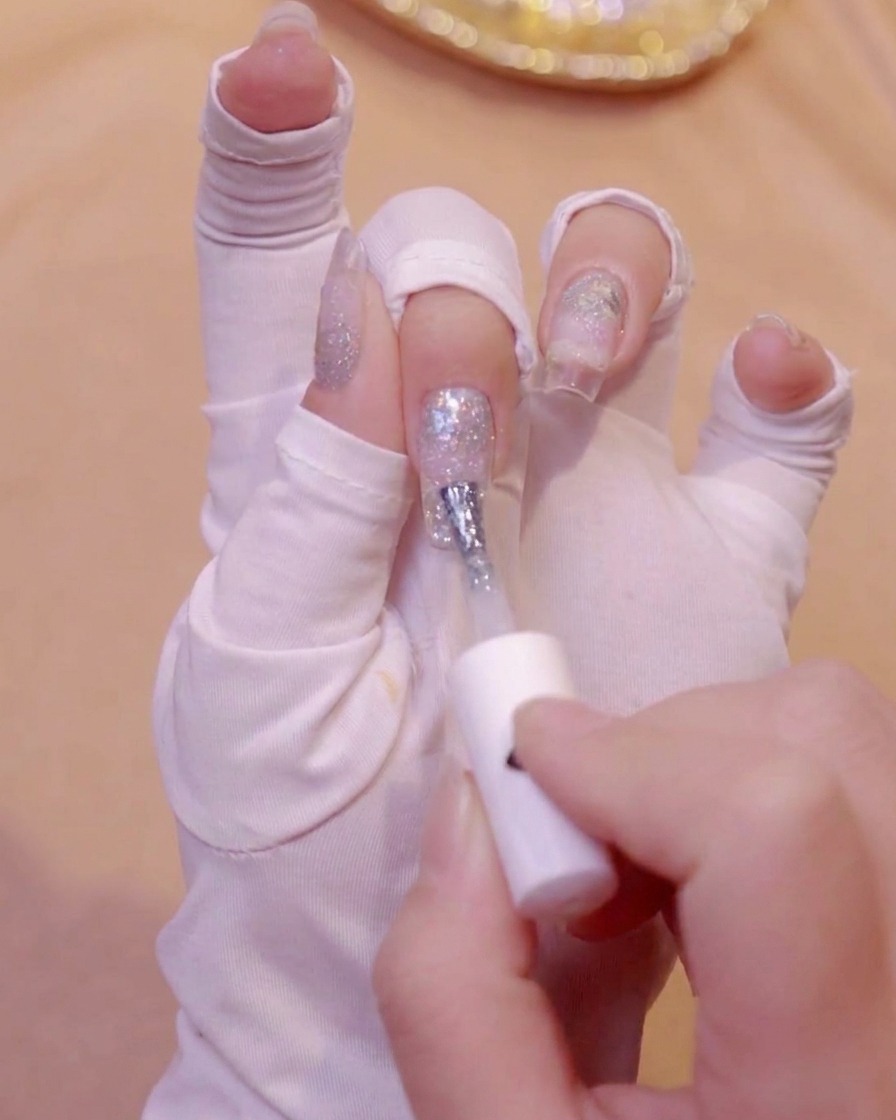Une main avec un gant blanc utilisant un pinceau pour appliquer du vernis gel blanc laiteux sur le bout dun ongle.