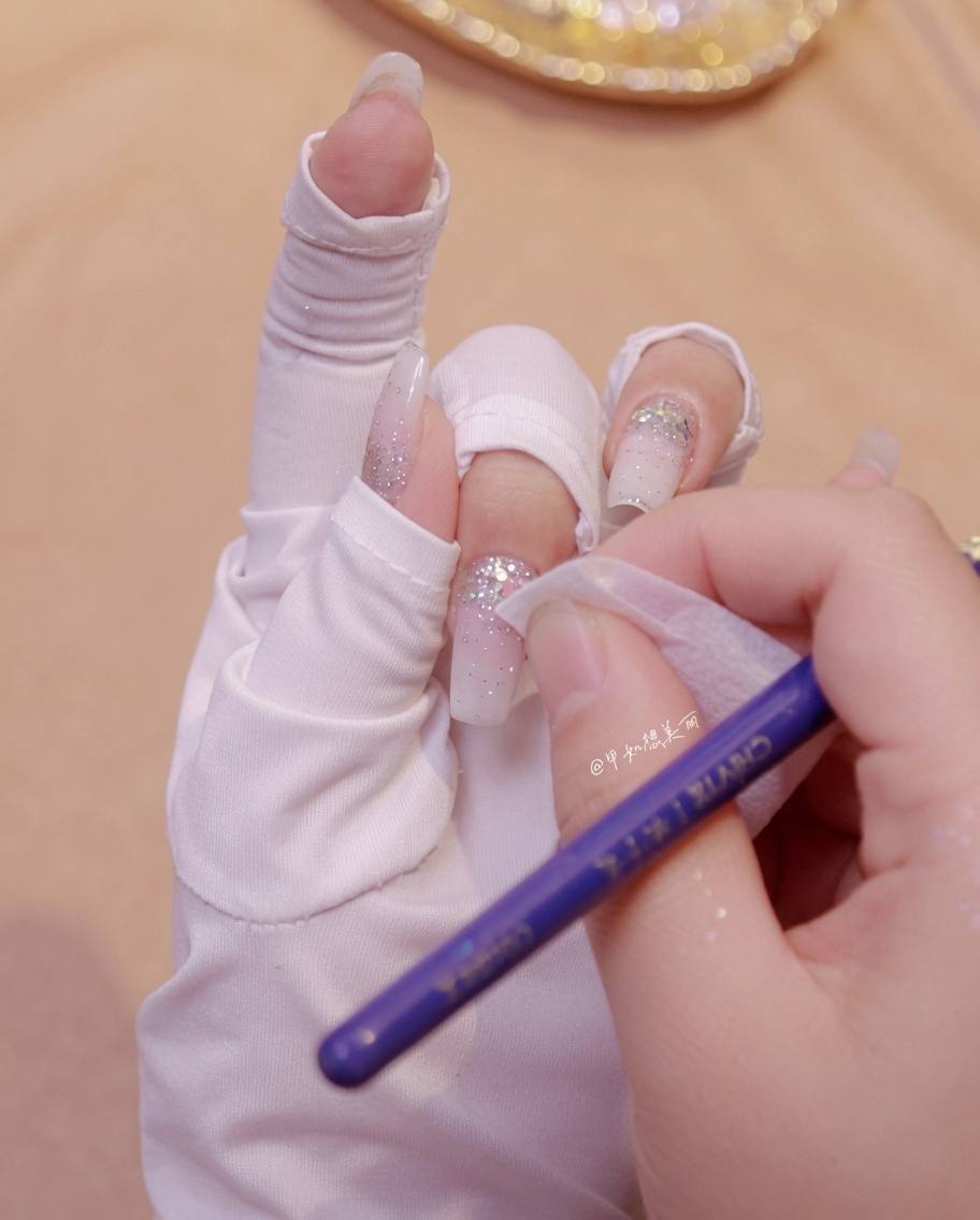 Une main utilisant un coton pour nettoyer autour des décorations de paillettes papillons sur un ongle manucuré.