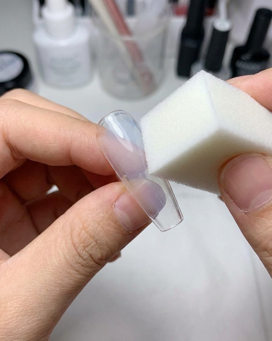 Une petite éponge à maquillage appliquant du gel bleu clair au centre dun faux ongle transparent.