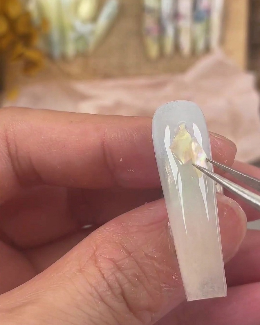 Une pince fine déposant des morceaux de coquillages irisés sur la surface humide du gel de longle.