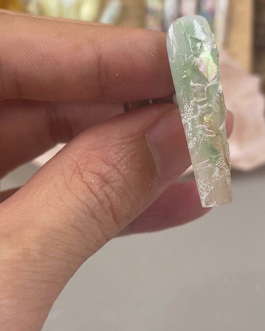 Gros plan dun ongle vert et transparent avec des éclats de coquillages irisés et des motifs de dentelle blanche estampés sur la surface.