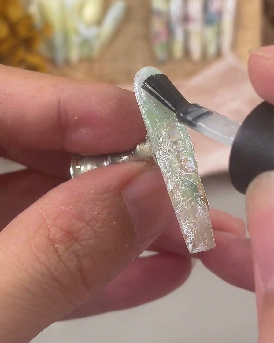 Un pinceau à ongles appliquant une couche épaisse de gel de construction transparent sur un ongle décoré de dentelle et de coquillages.