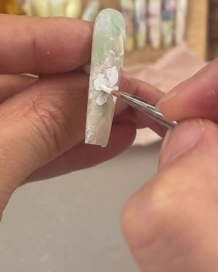 Un pinceau de détail fin appliquant du gel de peinture blanc pour dessiner une grande fleur sur un ongle mat vert et transparent.