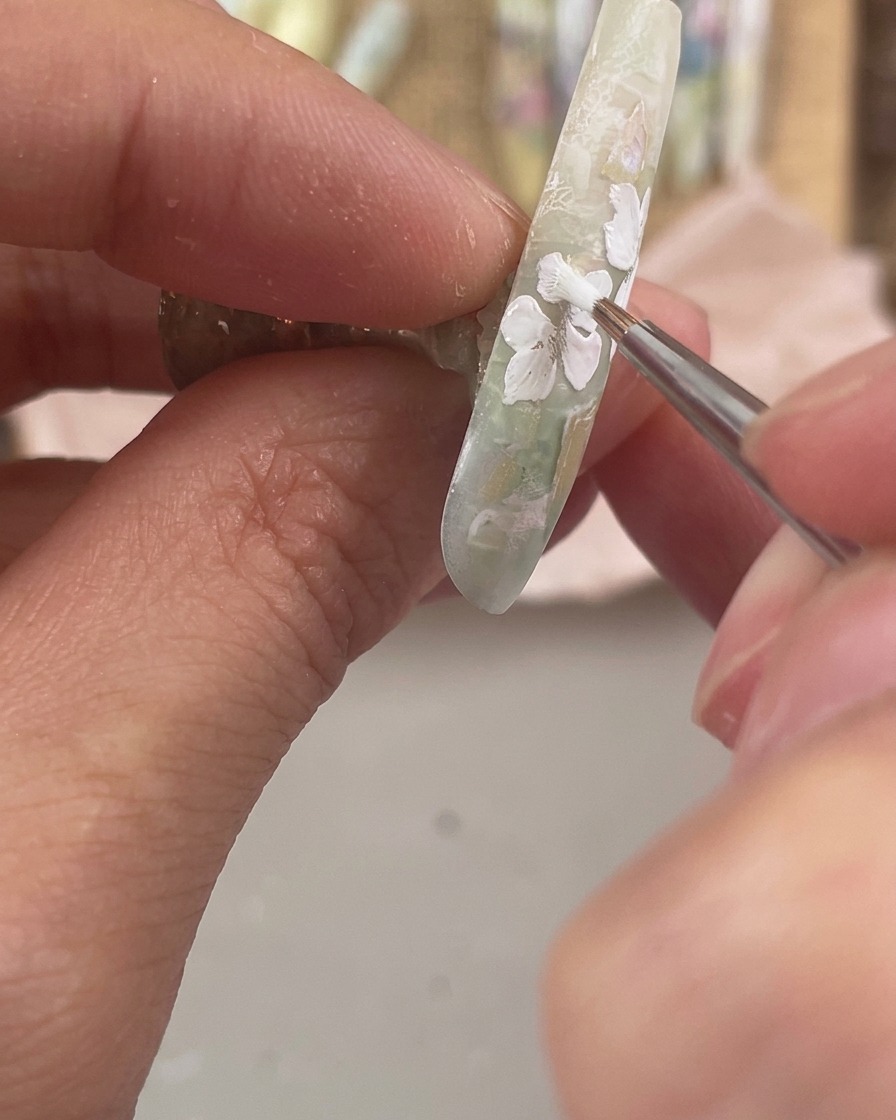 Un pinceau de détail fin peignant trois petits pétales floraux blancs le long du bord latéral inférieur dun ongle décoré.