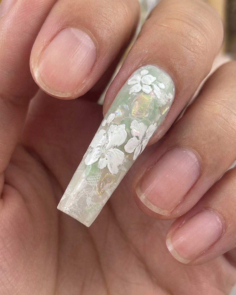 Gros plan dun ongle long sculpté montrant des fleurs en gel blanc terminées et de minuscules feuilles sur un fond vert avec des éclats de coquillages.