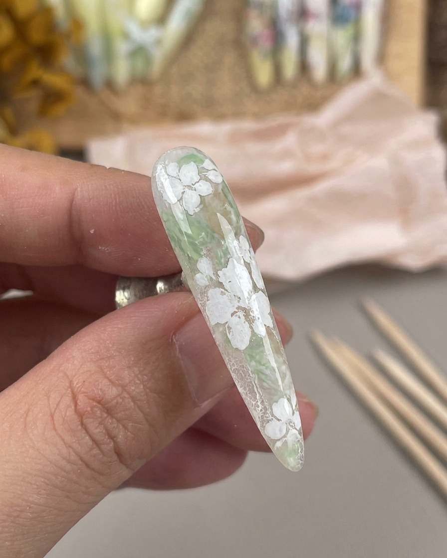 Un ongle long à la finition mate présentant un motif floral blanc, maintenu sur un support magnétique pour ongles.
