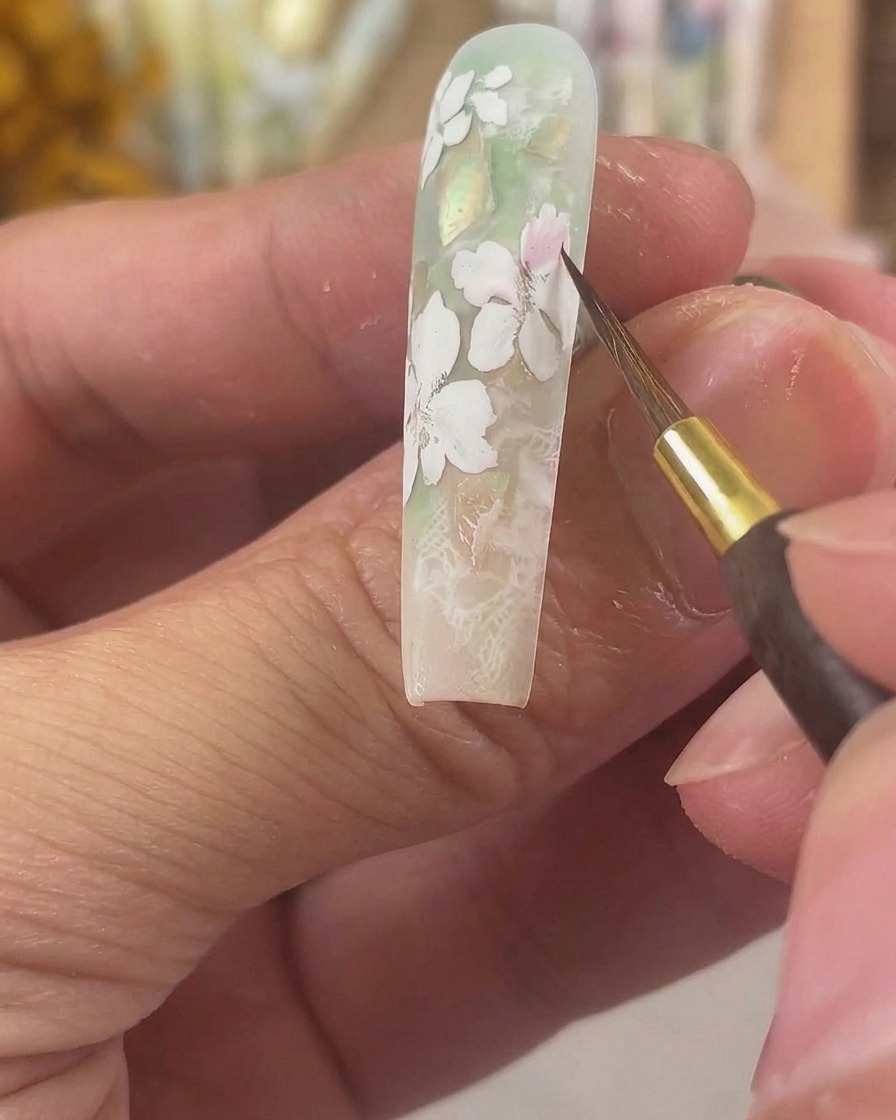 Un pinceau de détail fin appliquant un pigment aquarelle rose translucide sur des pétales en gel blanc mat dun ongle long sculpté.