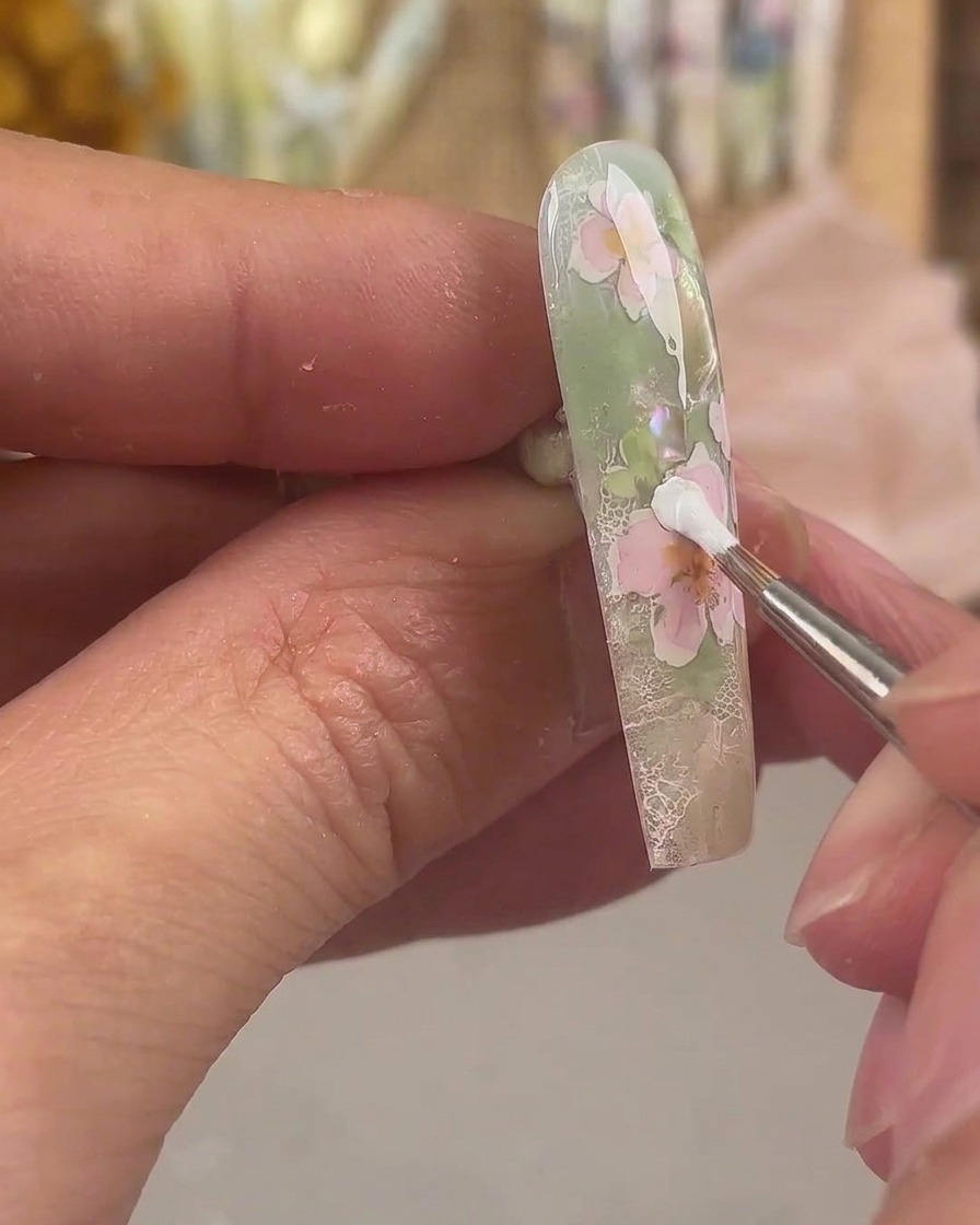 Un pinceau fin appliquant du gel de peinture blanc pour former une couche supplémentaire de pétales sur un motif de nail art floral rose.