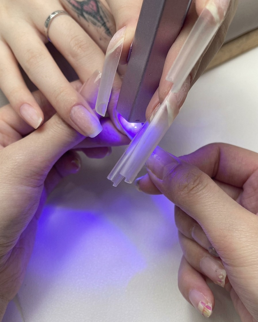 Gros plan dune main maintenant une capsule transparente sur un ongle naturel sous une petite lampe UV violette.