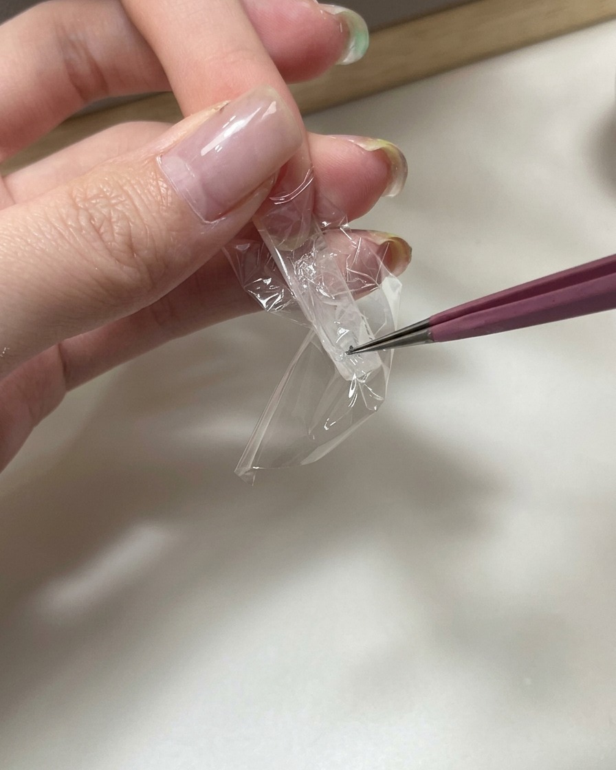 Une pince à épiler pressant un morceau de film plastique transparent sous une capsule pour créer une texture froissée.