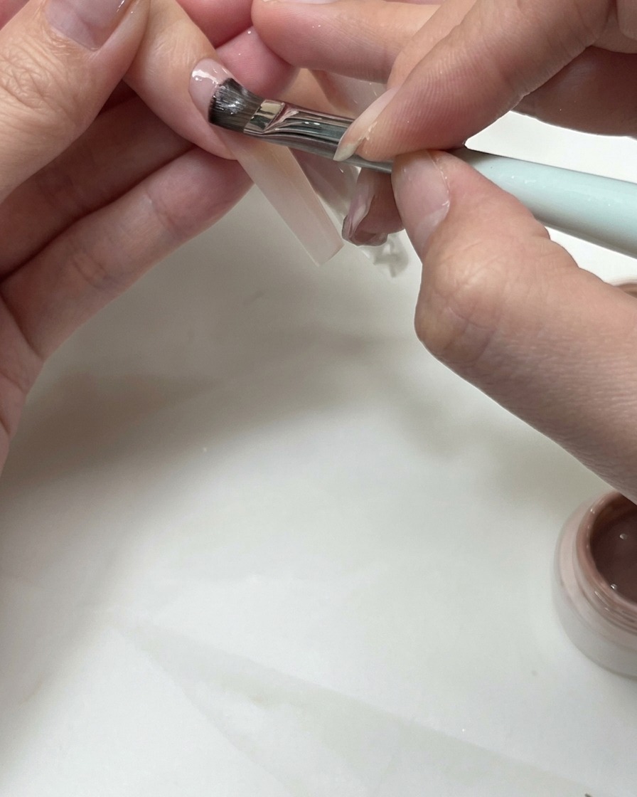 Un pinceau plat appliquant du gel nude pour sculpter une ligne de sourire de french sur un ongle.