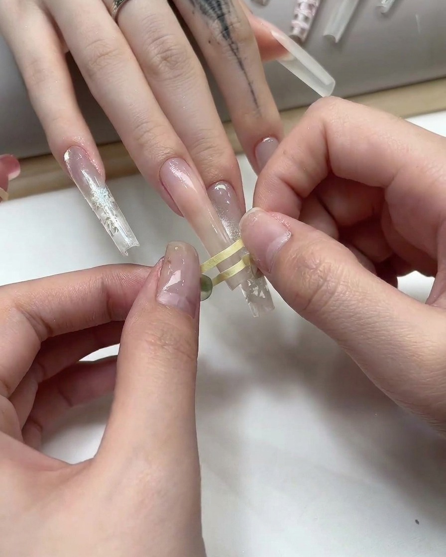 Des doigts plaçant un fin ruban de masquage doré en forme de grille sur un ongle au gel nude.