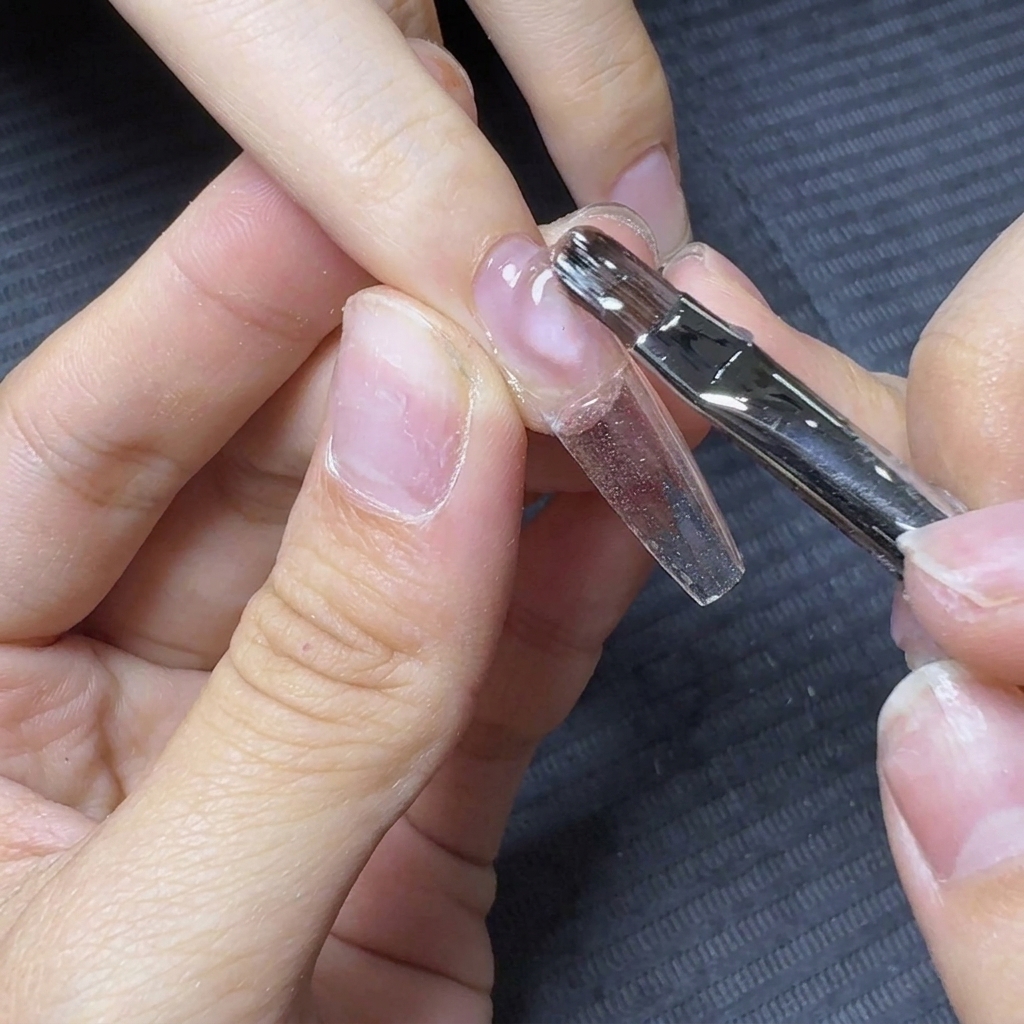 Un pinceau à ongles faisant flotter et étirant le gel de construction transparent sur toute la longueur de la capsule transparente.