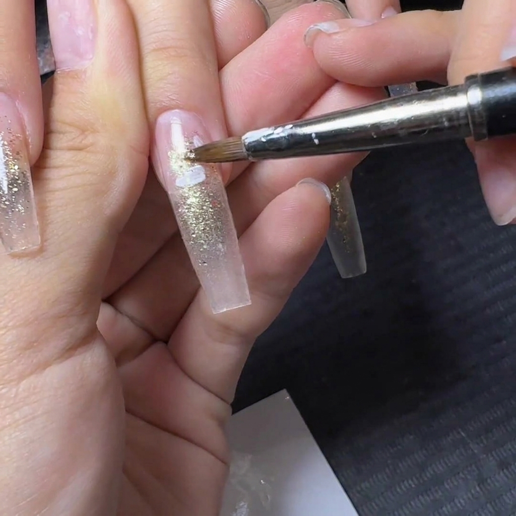 Un pinceau de détail fin appliquant des motifs de pétales de fleurs blancs sur un ongle pailleté translucide.