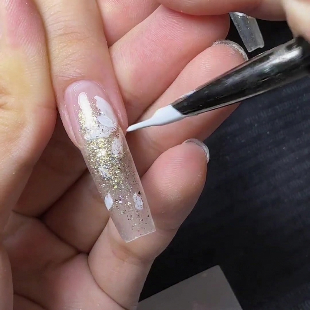 Un pinceau liner fin peignant de petits pétales de fleurs blancs sur un ongle transparent avec des paillettes dorées.