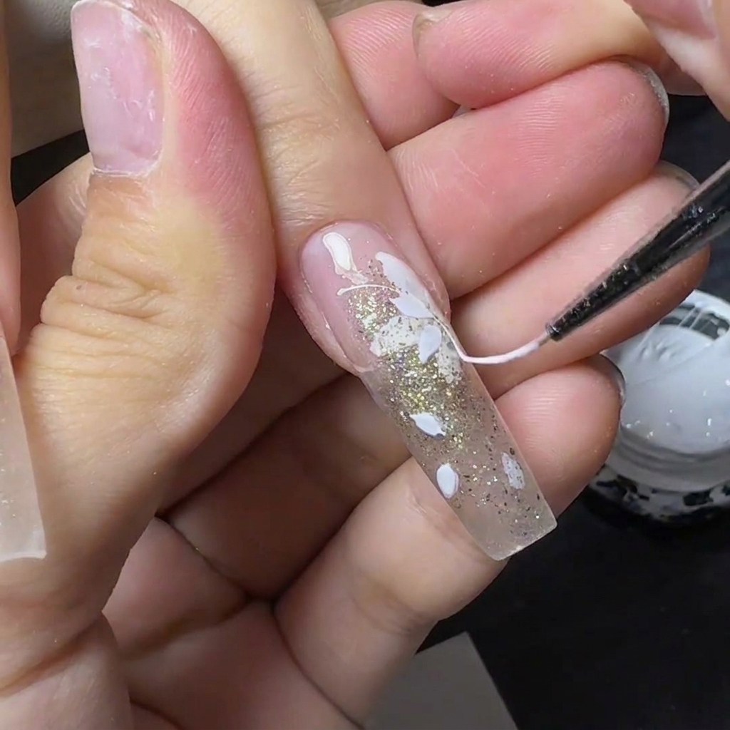 Un outil fin tirant du spider gel blanc filamenteux sur un ongle en gel décoré de fleurs blanches et de paillettes dorées.