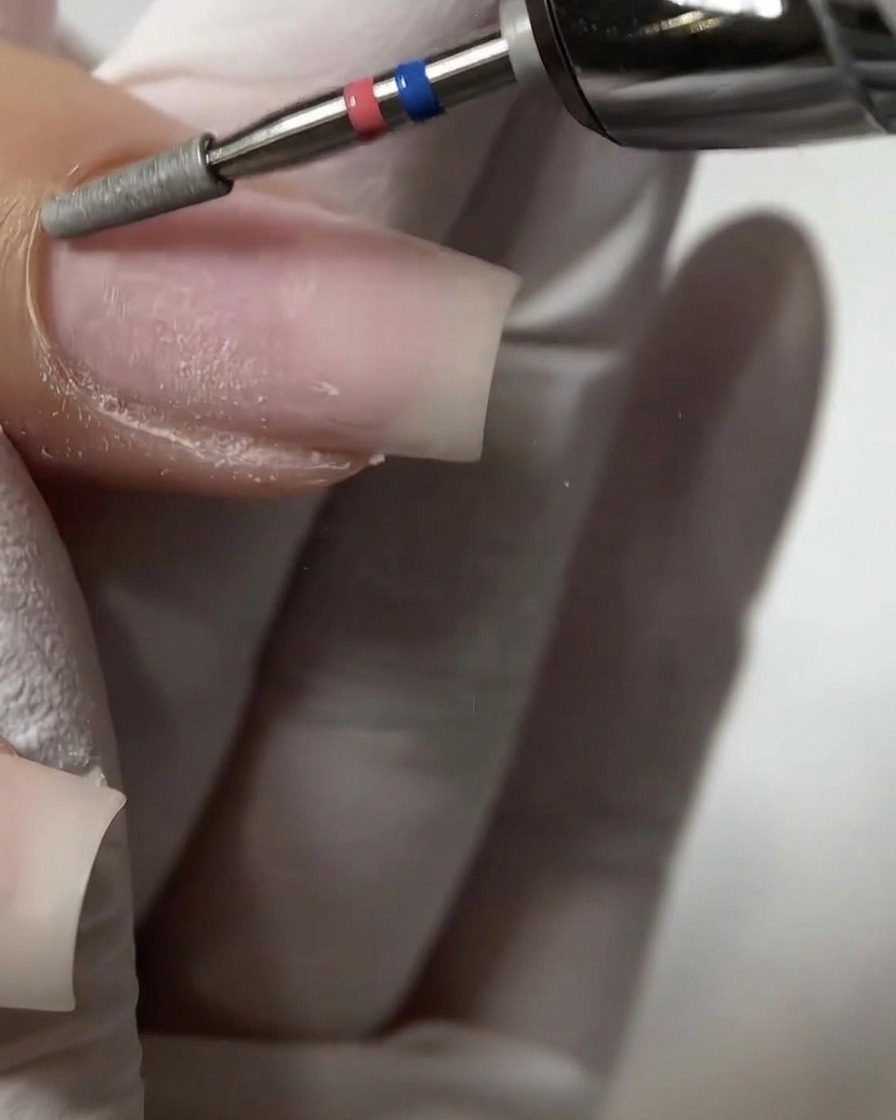 Gros plan sur un embout cylindrique plat de ponceuse exfoliant et repoussant délicatement le bourrelet proximal de la cuticule sur un ongle naturel.