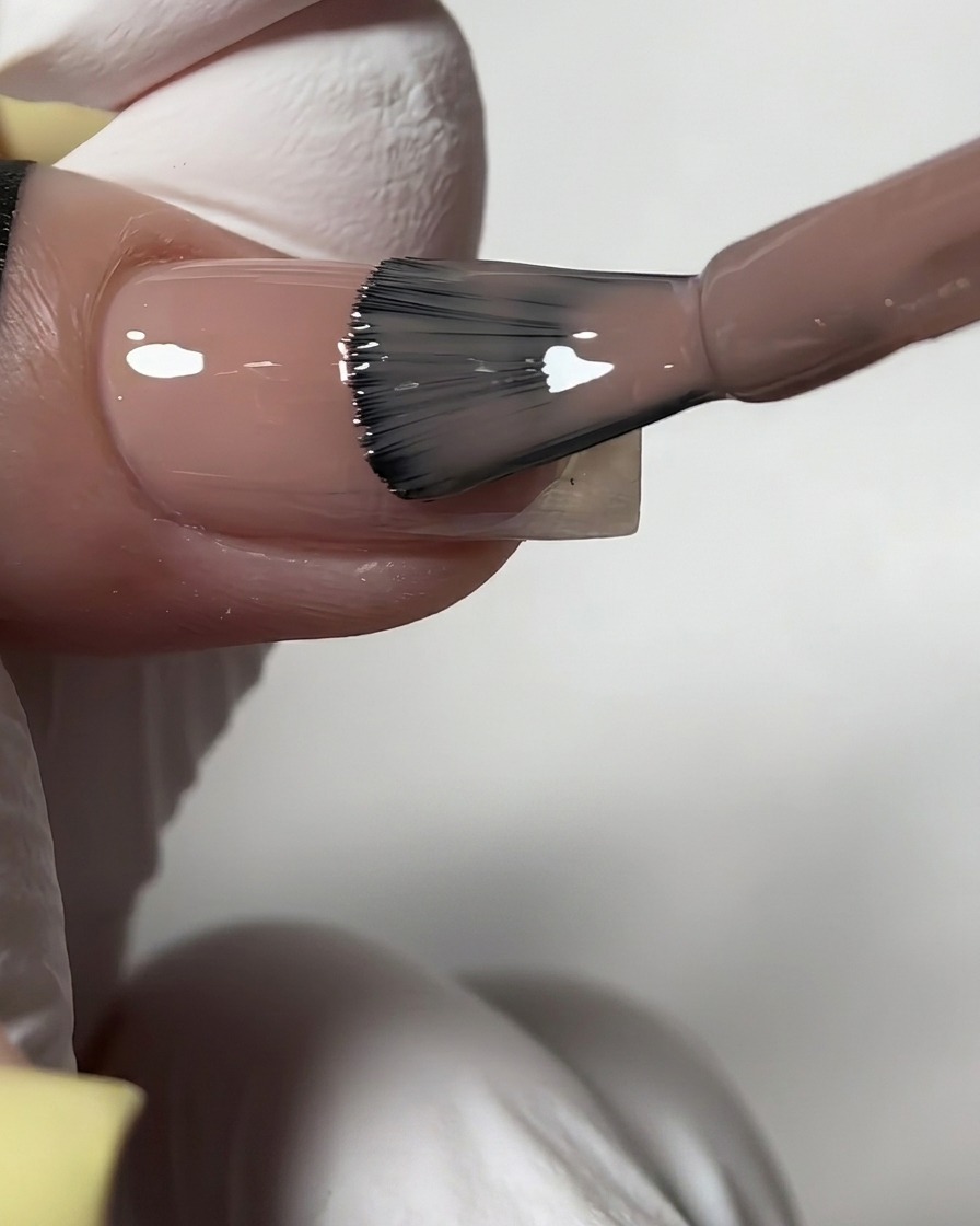 Un pinceau de flacon appliquant un vernis gel nude diaphane et brillant sur un ongle préparé.