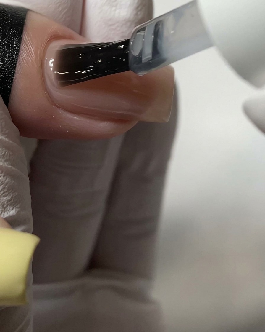 Un pinceau appliquant un top coat transparent sur un ongle en acrylique de couleur nude.