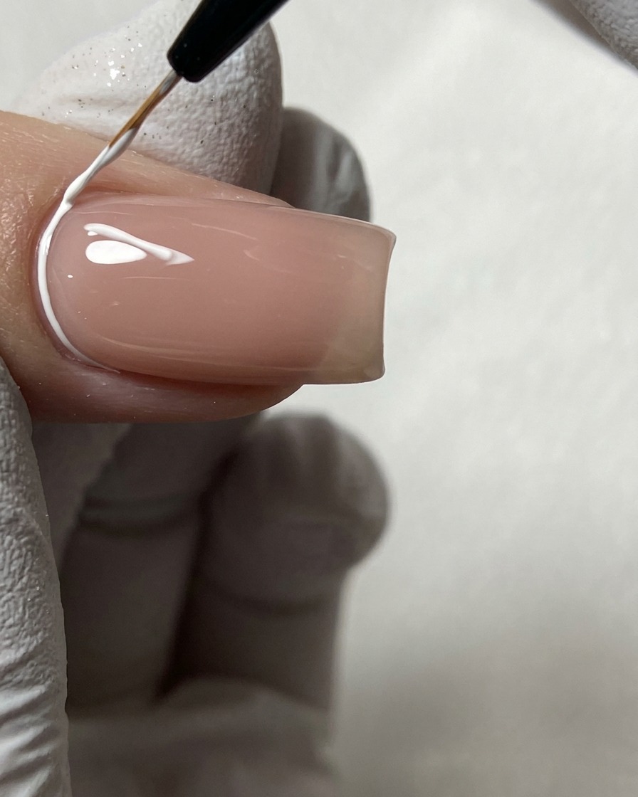 Un pinceau liner fin dessinant des lignes géométriques blanches près de la cuticule sur un ongle nude.