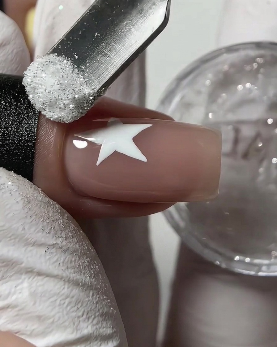 Un repousse-cuticules en métal saupoudrant de la poudre de paillettes de sucre blanche sur une étoile blanche humide tracée sur un ongle nude.