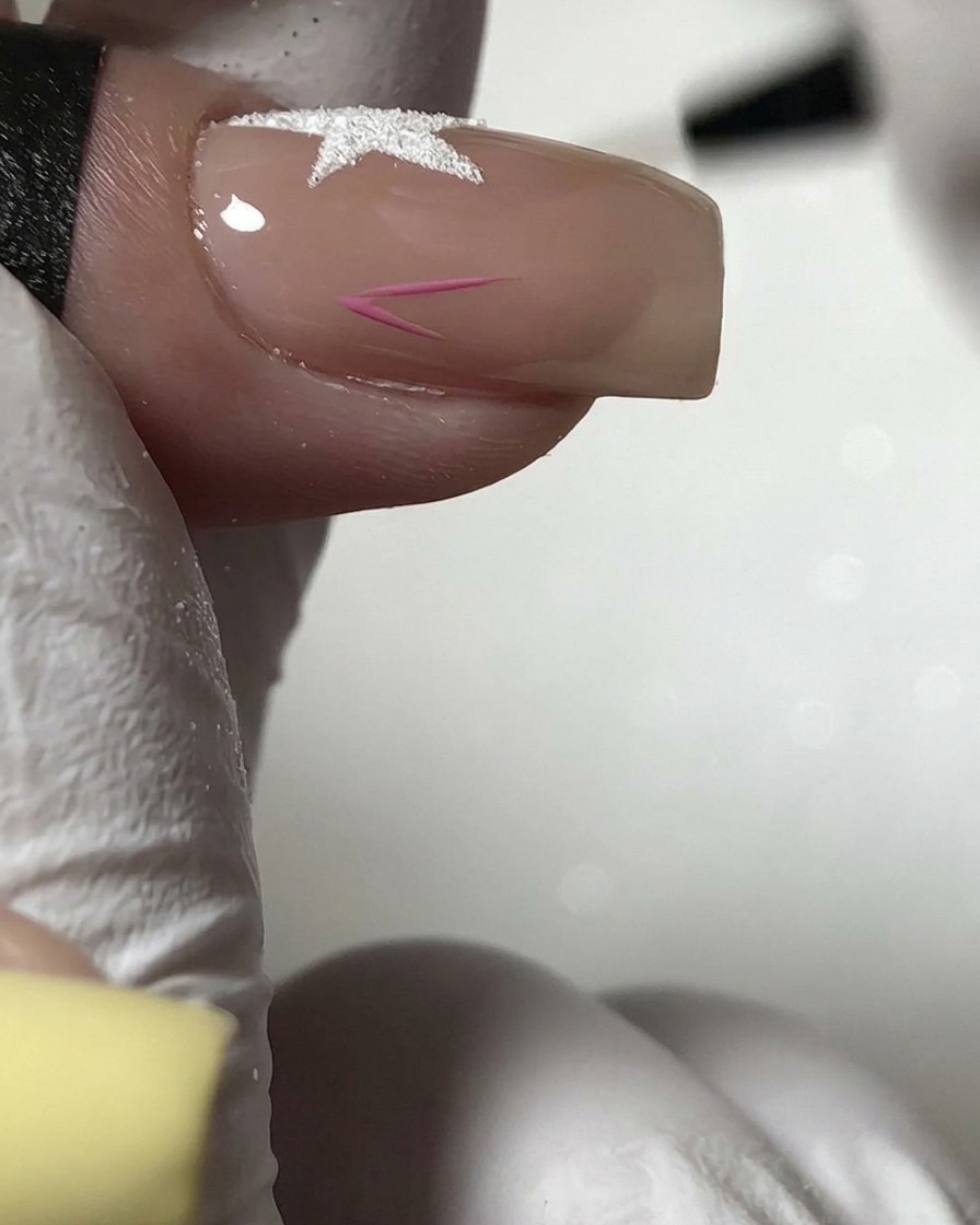 Un pinceau fin peignant le contour dune petite étoile rose sur la partie inférieure dun ongle nude.