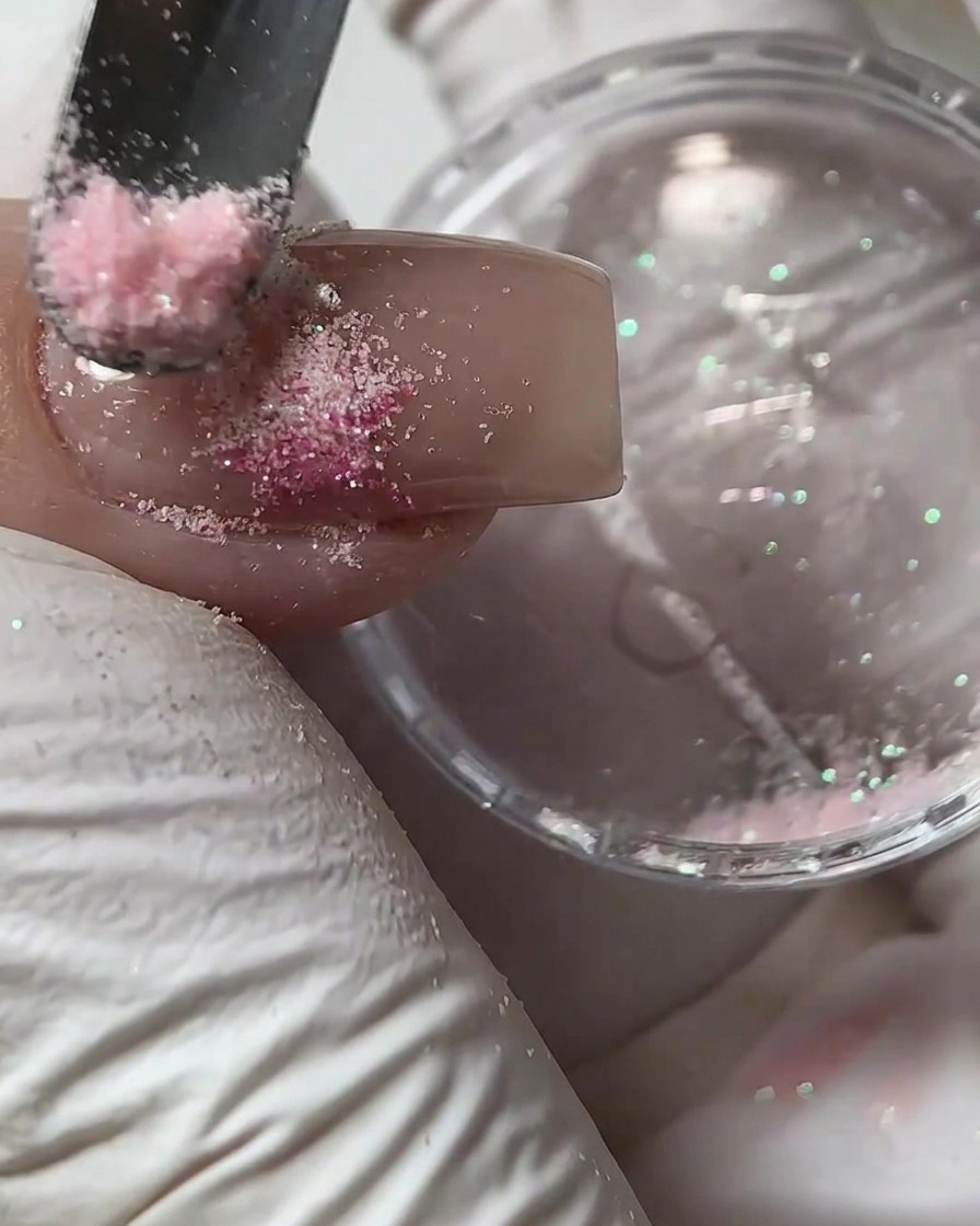Un petit outil dispersant de la poudre de sucre rose sur un motif en gel humide sur un ongle.
