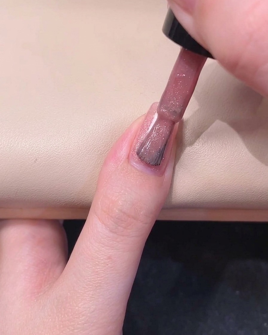Un pinceau à ongles peignant le côté droit dun ongle avec du vernis gel rose scintillant.