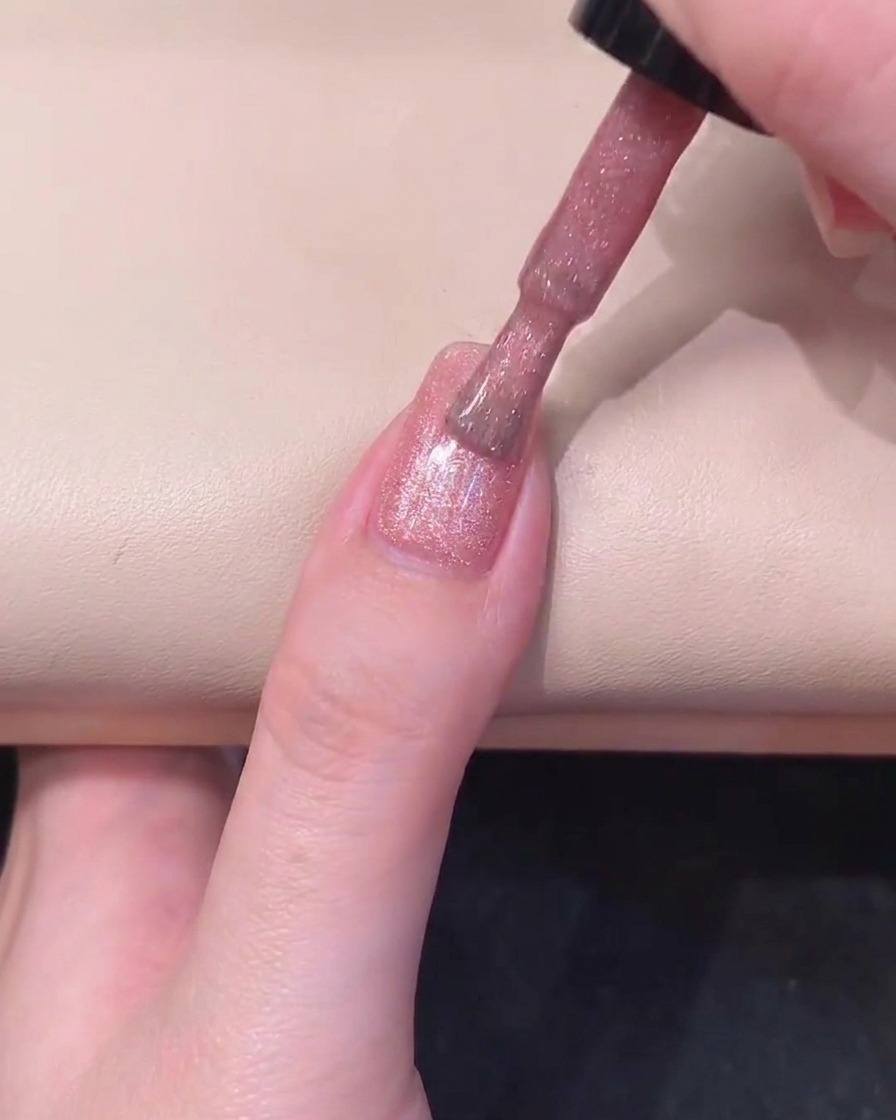 Un aimant cylindrique noir maintenu parallèlement au côté droit dun ongle enduit de gel rose pour activer le scintillement.