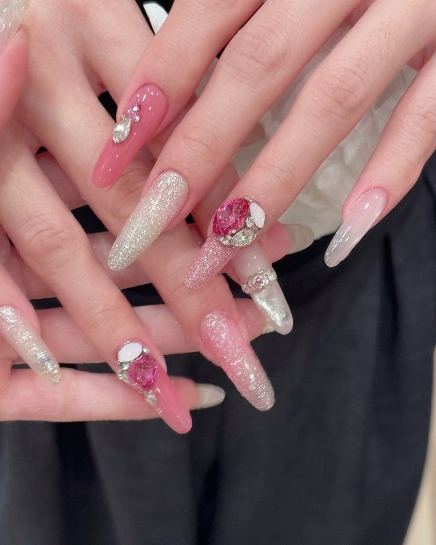 Deux mains parfaitement manucurées reposant sur un tissu sombre, arborant de longs ongles en amande roses avec des dégradés de paillettes argentées et de gros strass en forme de cœur.