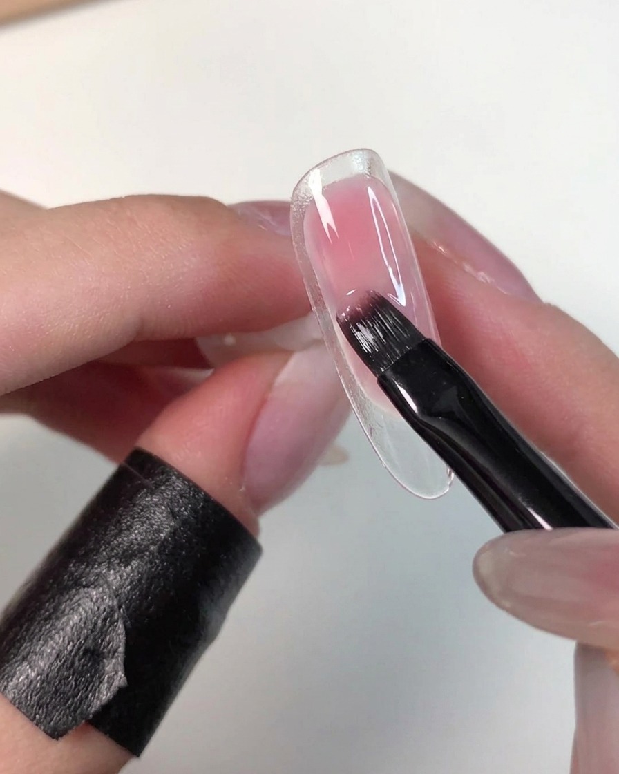 Une main tenant une capsule transparente pendant quun pinceau plat estompe doucement le gel rose humide pour adoucir ses bords.