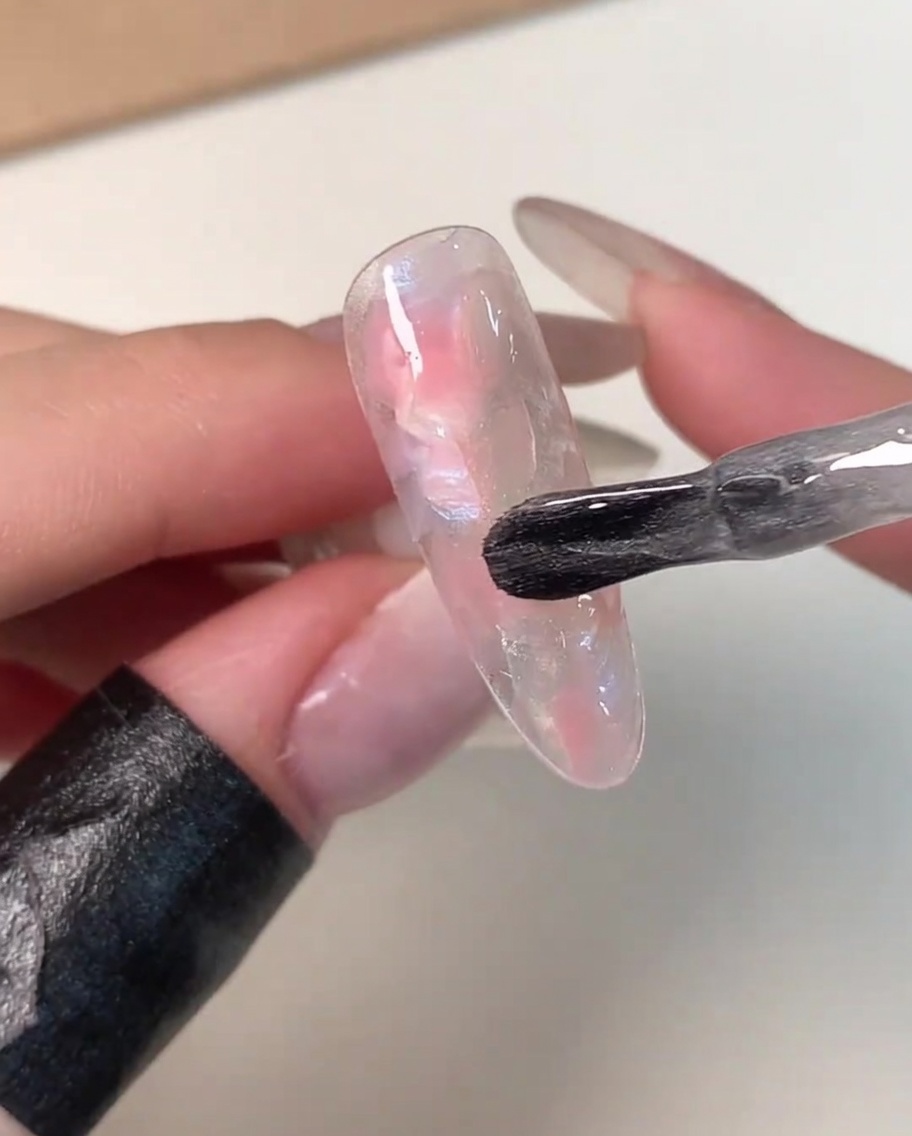 Un pinceau de vernis appliquant une couche de gel magnétique transparent et nacré sur une capsule marbrée rose et blanche.