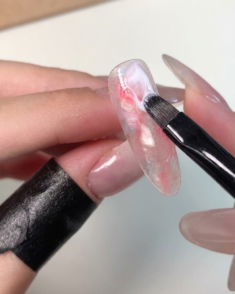 Un petit pinceau à gel appliquant un trait de gel blanc laiteux près de la cuticule dune capsule marbrée pour affiner le motif.