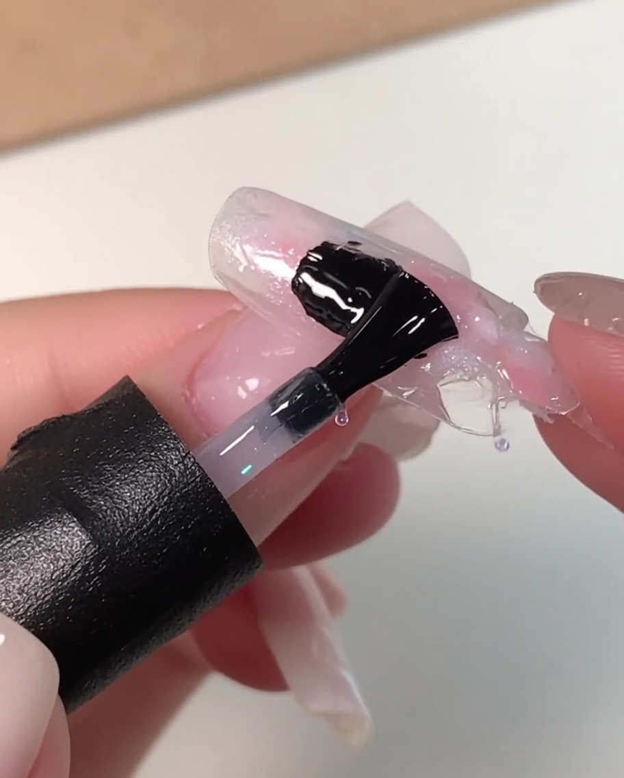 Un pinceau à ongles noir appliquant une généreuse couche de gel dégalisation transparent sur un motif marbré rose, blanc et bleu.