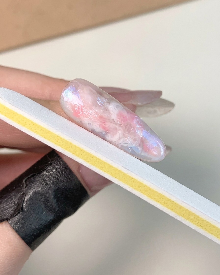 Une main tenant une lime à ongles blanche et jaune, polissant la surface dun ongle en gel marbré, brillant et catalysé.