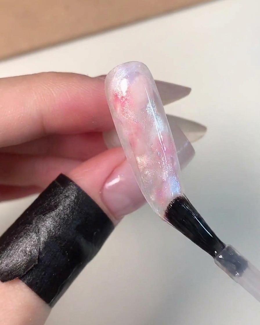 Un pinceau appliquant un top coat transparent et brillant sur un ongle effet boule de verre marbré rose, bleu et blanc magnifiquement fini.