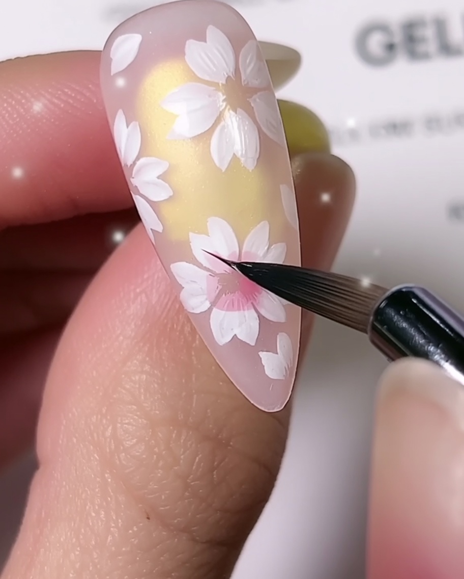 Un gros plan montrant un pinceau de détail fin ajoutant un ombrage rose au centre des pétales de cerisier blancs sur un ongle en amandecite: 1.