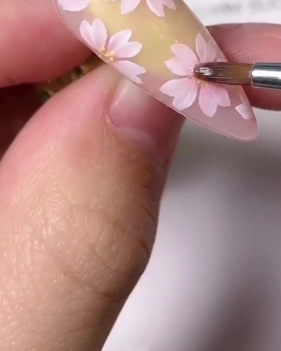 Un gros plan dun pinceau de détail fin appliquant de petits accents dorés au centre dun cerisier rose peint sur un ongle.