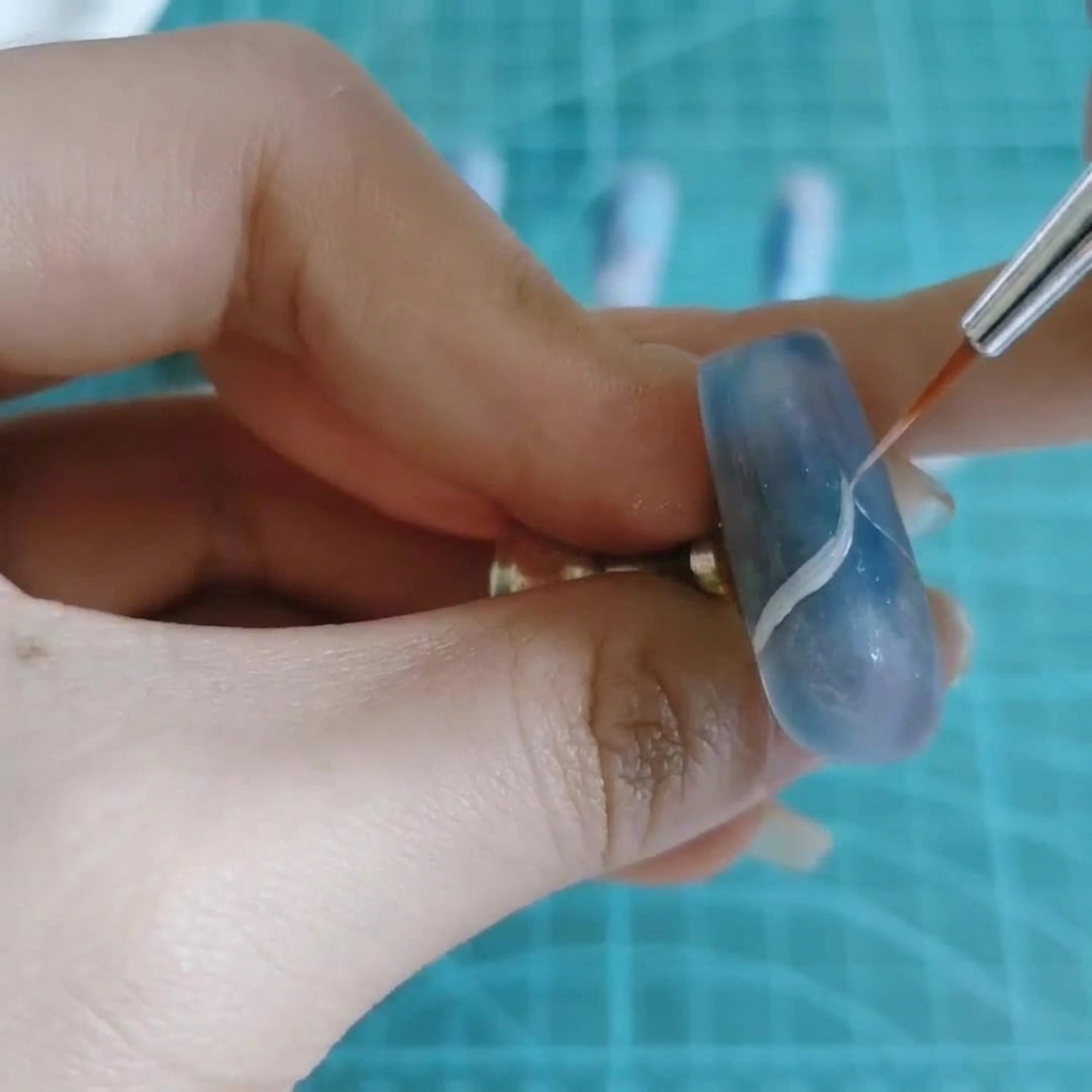 Un pinceau fin pour nail art dessinant une fine ligne blanche ondulée sur un ongle marbré bleu et violet.