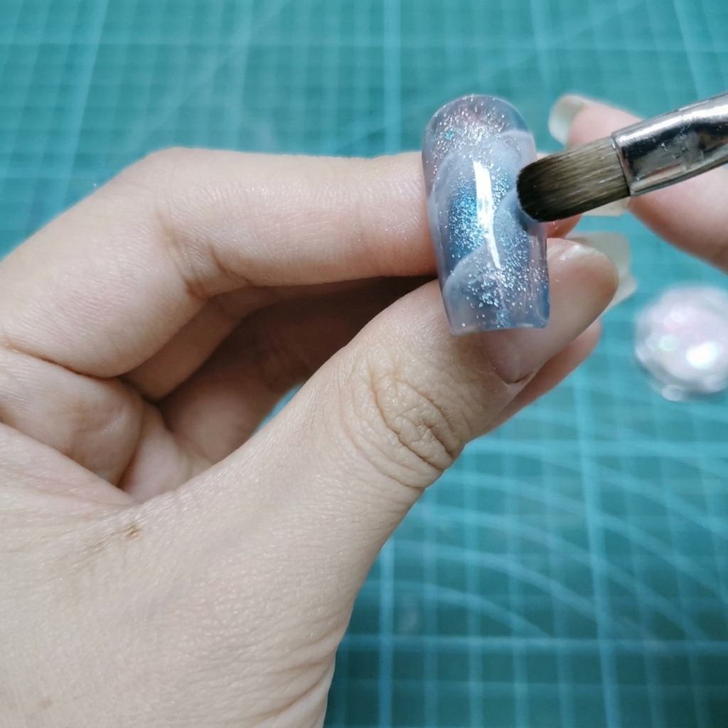 Un pinceau plat de maquillage tapotant des particules de paillettes holographiques sur la surface humide dun ongle en gel.